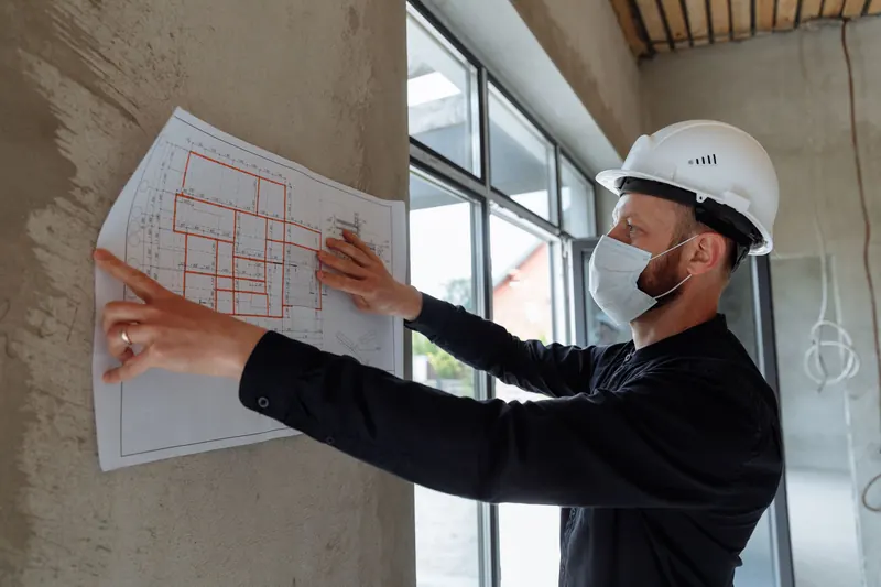Chef de chantier masqué et casqué consultant le plan d'un bâtiment affiché au mur