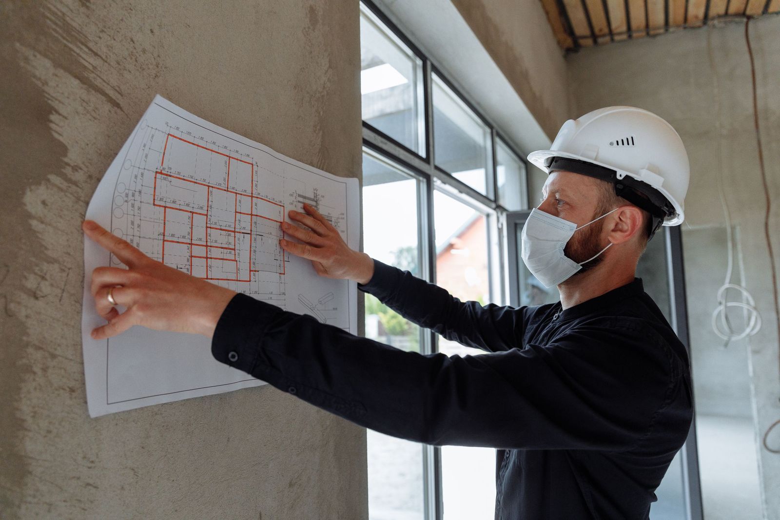 Chef de chantier masqué et casqué consultant le plan d'un bâtiment affiché au mur